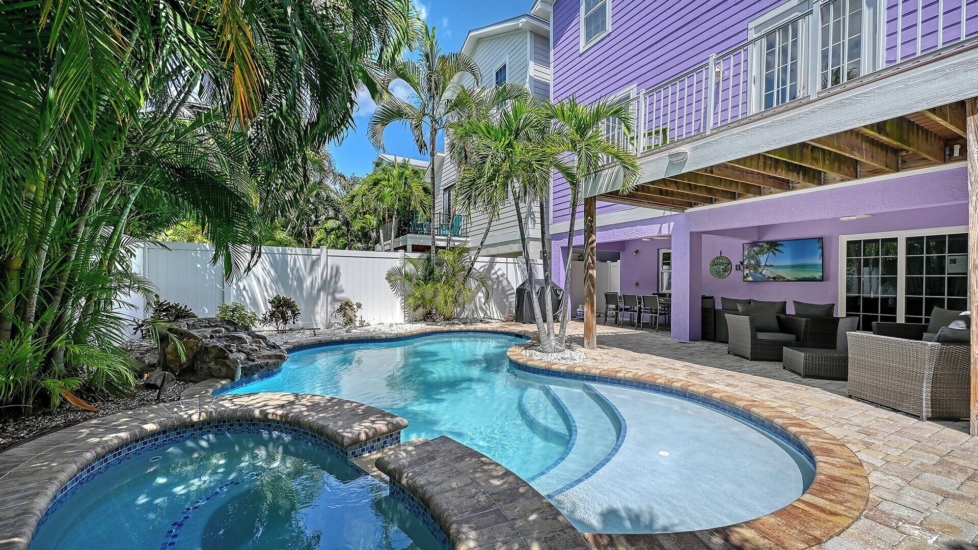 A swimming pool with a curved design, surrounded by tropical plants and a white fence.