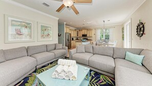 Ein Wohnzimmer mit zwei grauen Sofas, einem Couchtisch mit Handtüchern, einem Deckenventilator und einer Küche im Hintergrund.
