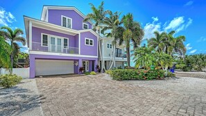A two-story house with a purple exterior, a white garage door, and a paved driveway. - Casa Morado | Luxury Holmes Beach Retreat with Heated Pool and Hot tub, Steps to Beach  I .PET FRIENDLY. ANNA MARIA ISLAND! (Holmes Beach)