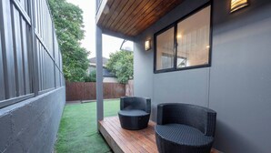 Een terras met twee rieten stoelen, een houten dek en een grijze muur met een raam.