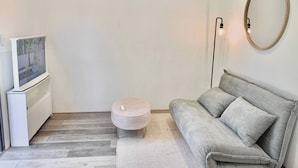 Een moderne woonkamer met een grijze bank, een rond hockerkussen, een flatscreen televisie en een ronde spiegel aan de muur.