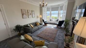 A living room with a sofa set, a coffee table, a rug, and a fireplace. There are potted plants and a large window with a view.