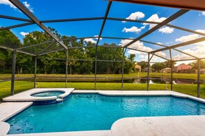 A swimming pool with a hot tub, surrounded by a covered area with a metal frame and glass walls, set against a backdrop of trees and a clear sky. - Whispering Waters Villa | 4BR/2BA Davenport, Water View (Davenport)