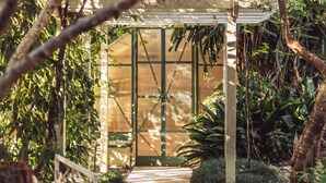 A glass-enclosed structure with a wooden frame, surrounded by dense foliage.