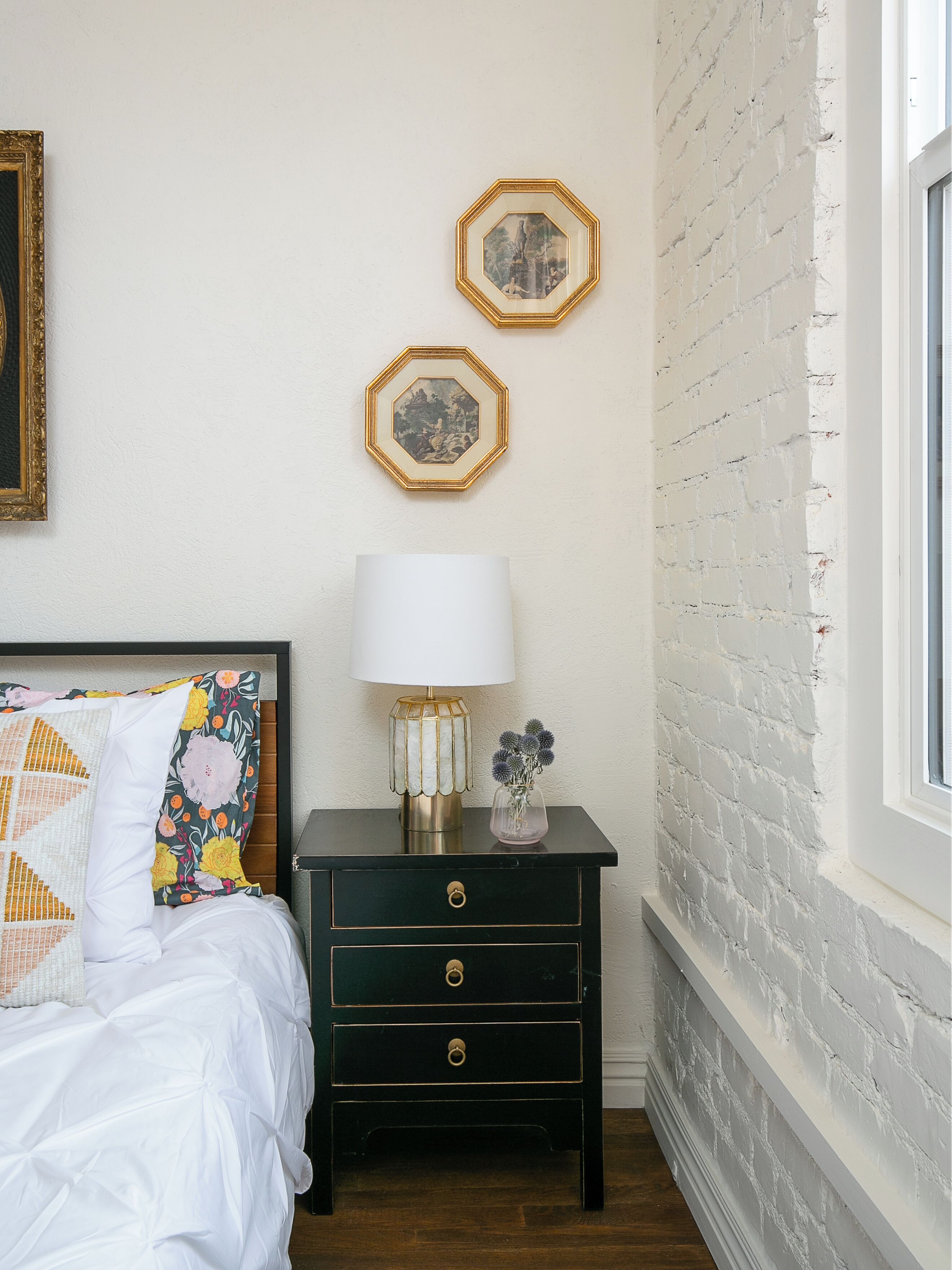 A bedroom with a bed, a nightstand with a lamp, two framed pictures on the wall, and a brick wall.