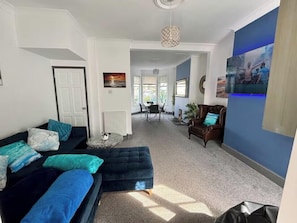 A modern living room with a sofa, a coffee table, and a dining area in the background. - Coastal House opposite beach sleeps 6 Pet friendly (Southend-on-Sea)