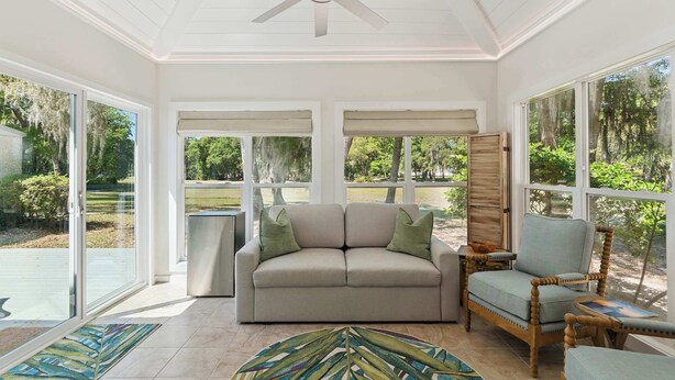 Ein Wintergarten mit Deckenventilator, einem beigefarbenen Sofa mit grünen Kissen, einem Holzstuhl und einem gemusterten Teppich.
