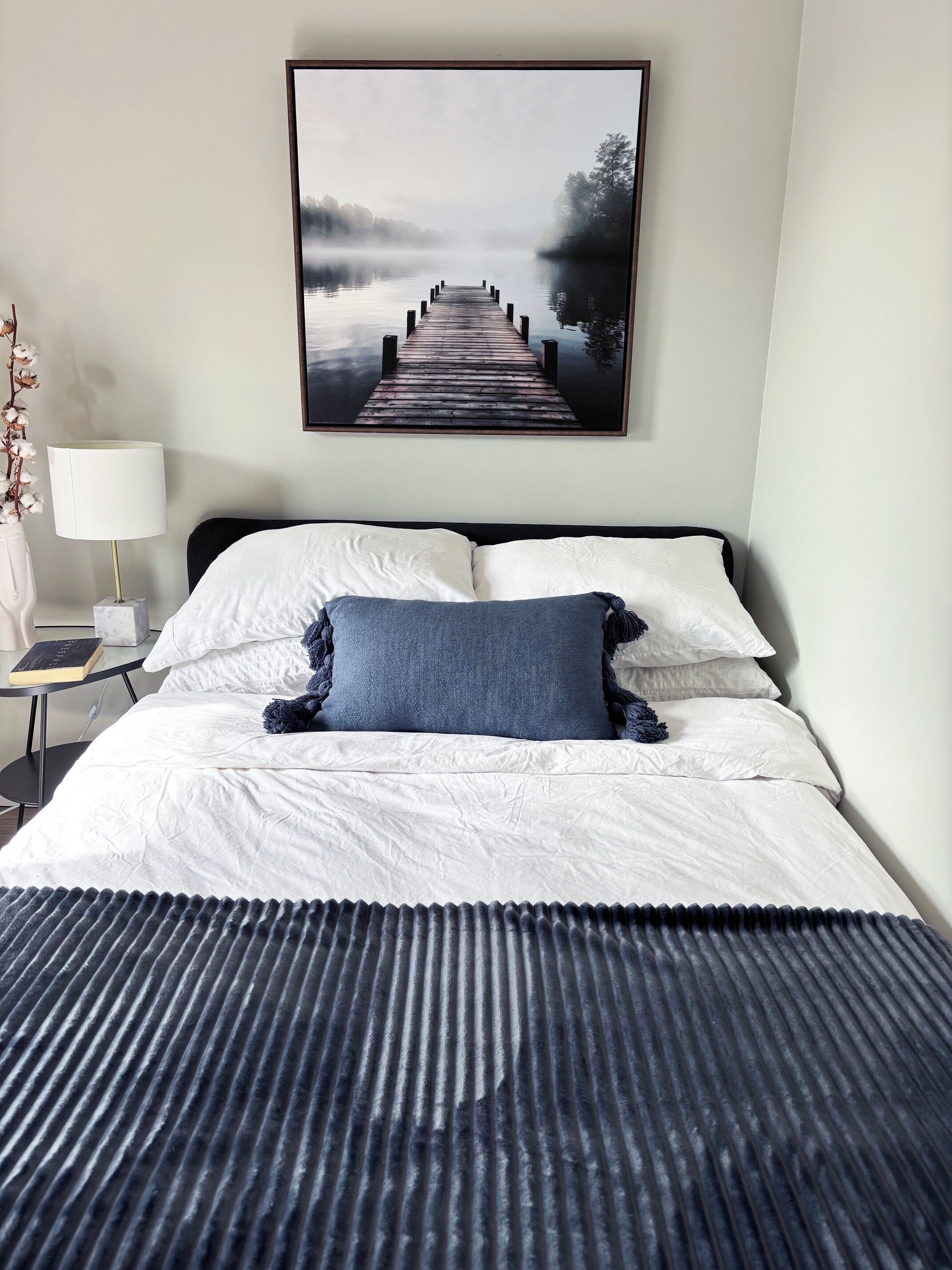 A neatly made bed with white linens and a dark blue throw blanket, a framed picture of a pier on the wall, and a bedside table with a lamp and a book.