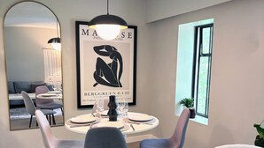 Een eethoek met een ronde tafel, vier stoelen, een hanglamp, een lijstwerk, een raam en een spiegel.