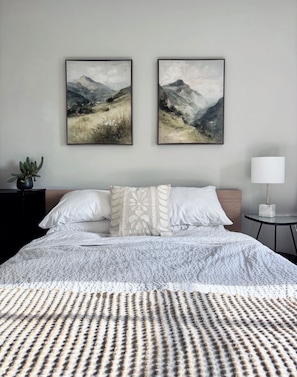 Une chambre à coucher avec un lit, deux tableaux encadrés, une table de chevet avec une lampe et une plante en pot.