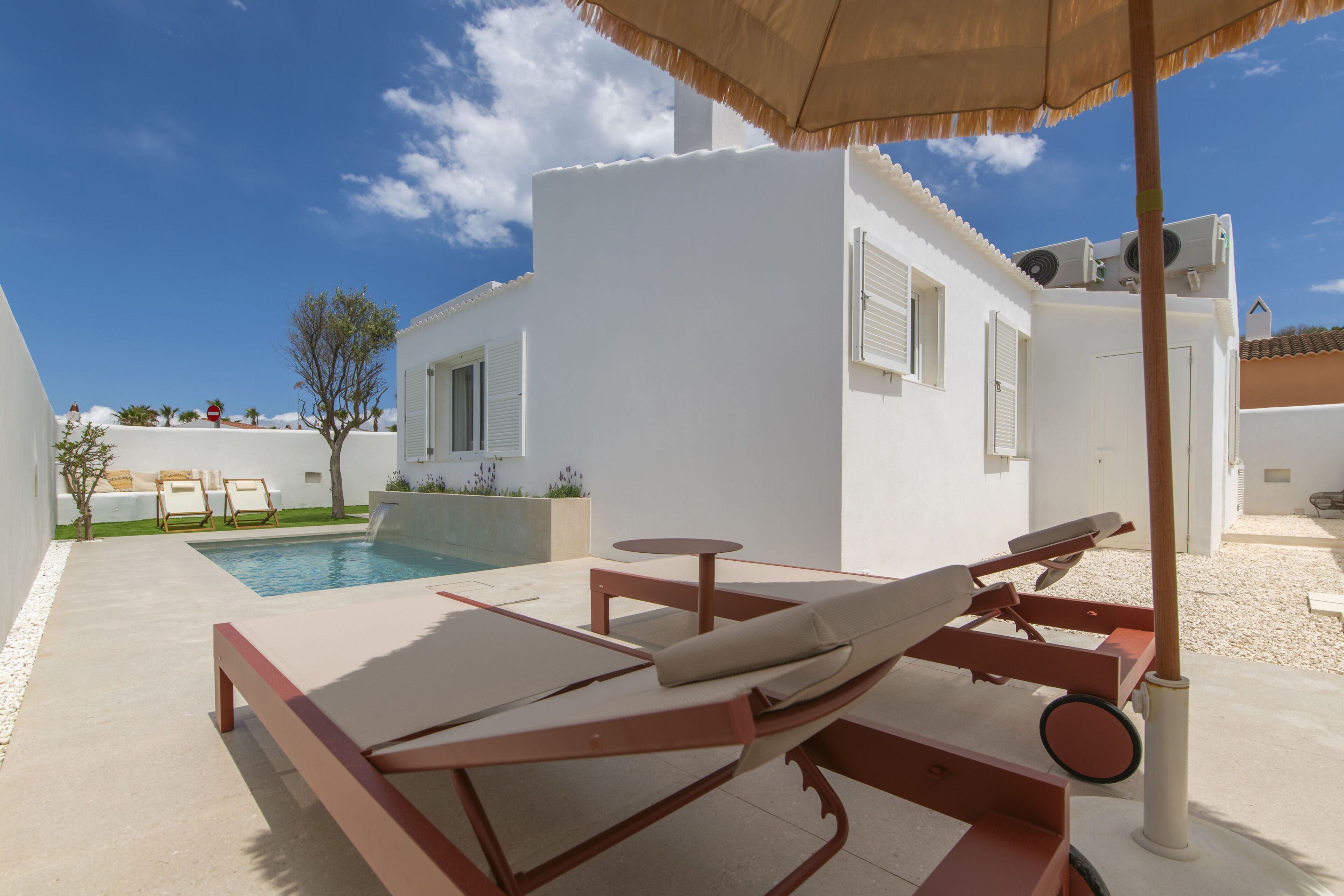 A white villa with a pool, lounge chairs, and a table under a striped umbrella.