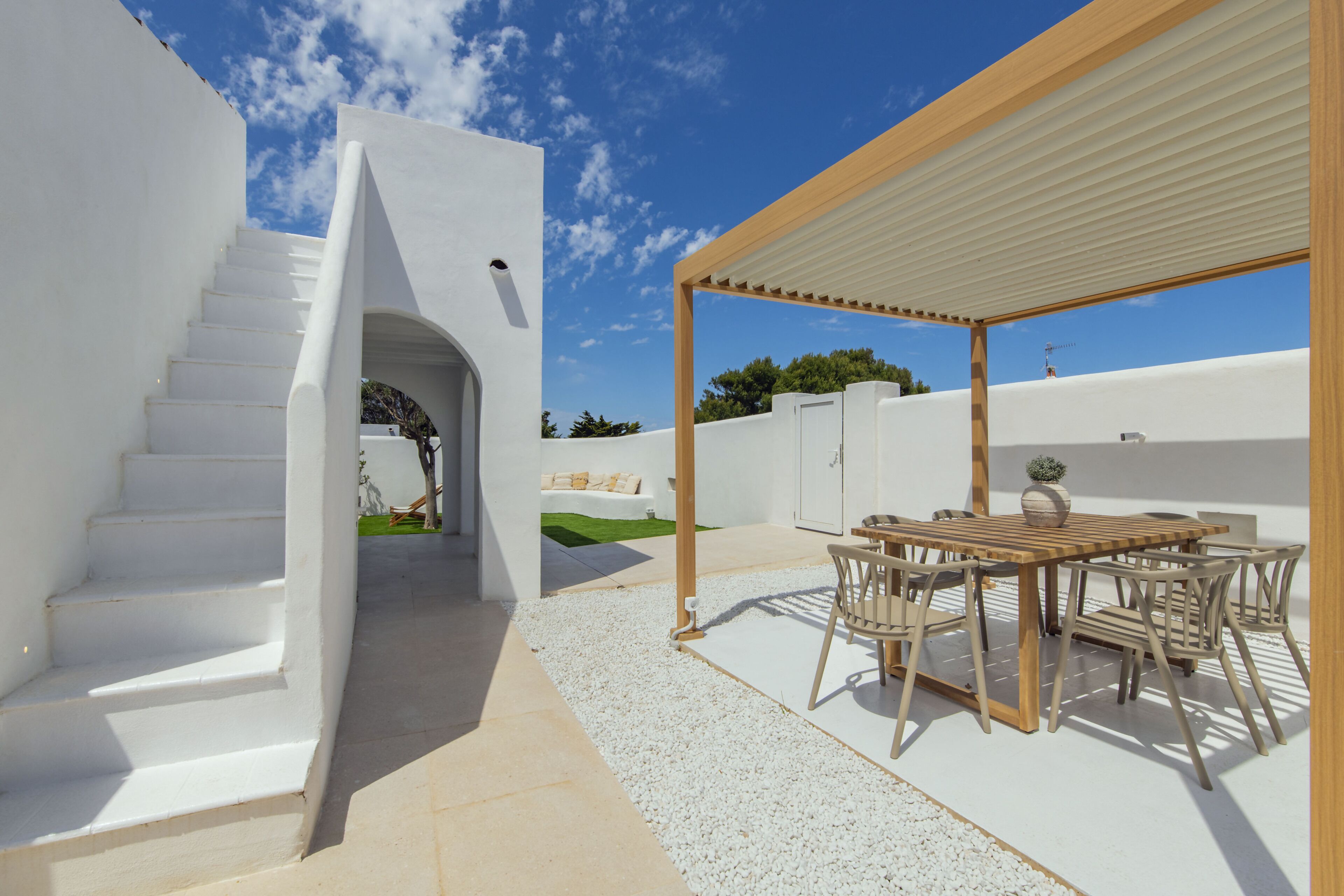 A modern outdoor patio with a wooden table and chairs, a white staircase, and a covered seating area.