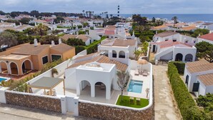 Zona residencial costeira com edifícios brancos, piscina e um farol ao fundo.