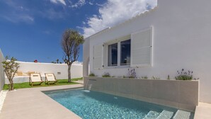 Piscina clara com borda azulejada, duas espreguiçadeiras, uma pequena árvore e um edifício branco com persianas.