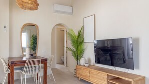 Uma sala de jantar com mesa de madeira, cadeiras brancas, um espelho, um vaso com planta, uma televisão sobre suporte de madeira e um aparelho de ar condicionado.