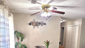 Ein Essbereich mit einem Glastisch, vier Stühlen, einem Deckenventilator und einem Wandbild.