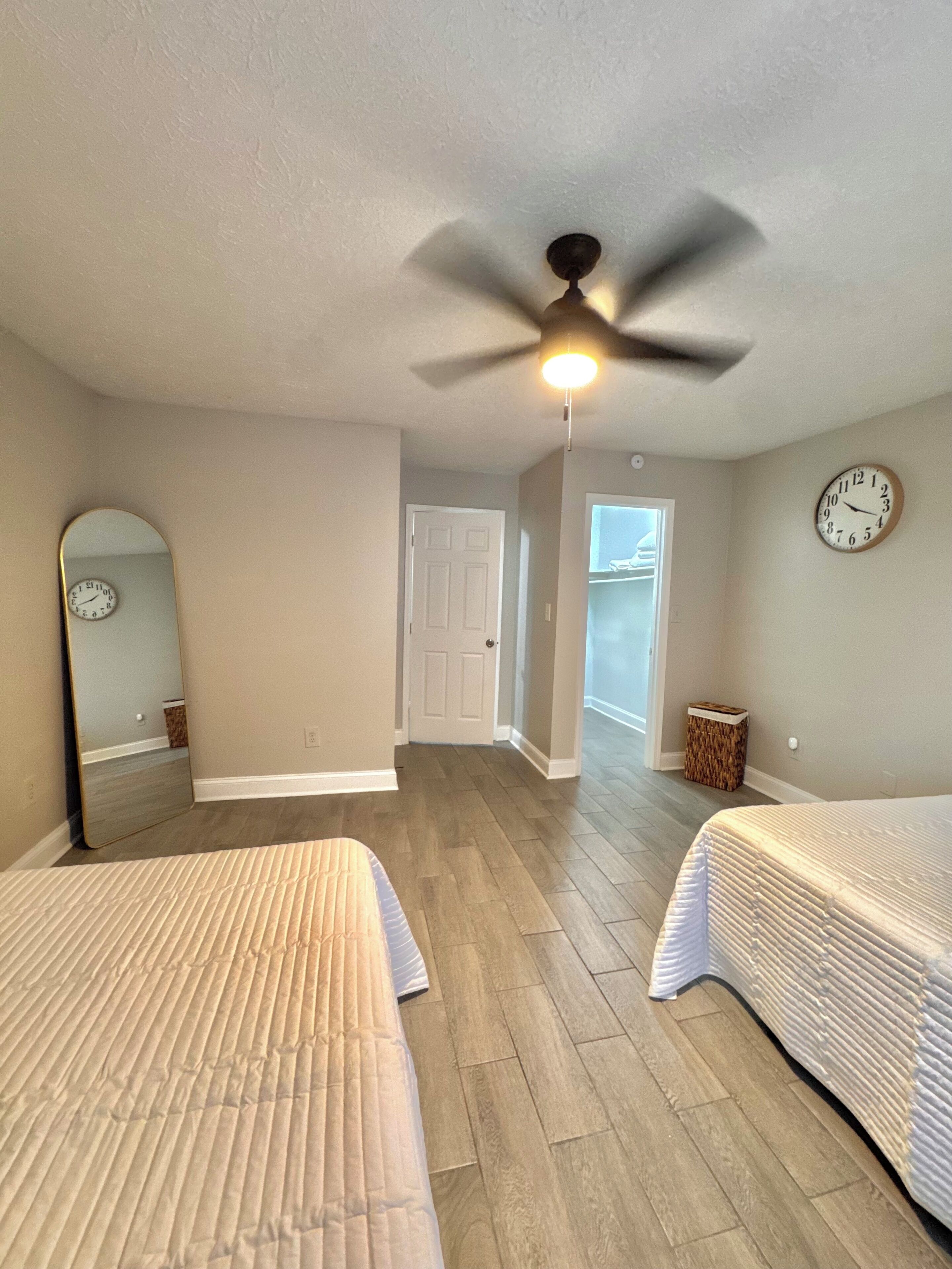 A bedroom with two beds, a ceiling fan, a clock, and a door leading to another room.