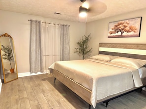 Une chambre à coucher avec un lit, un ventilateur de plafond, une fenêtre avec des rideaux, un tableau accroché au mur et un miroir agrémenté d’une plante.