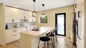 A modern kitchen with white cabinets, a central island, bar stools, pendant lighting, and a view of the outdoors through a glass door.