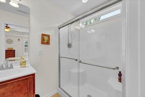 A bathroom with a glass shower enclosure, a white sink with a wooden vanity, a mirror, and a clock on the wall.