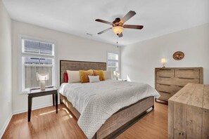 A bedroom with a bed, a wooden dresser, a nightstand, a ceiling fan, and a window with blinds.