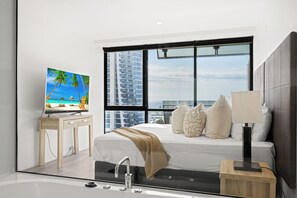 Ein modernes Hotelzimmer mit großem Fenster, einem Bett mit Kissen, einem Nachttisch mit Lampe und einem Fernseher auf einem Schreibtisch.