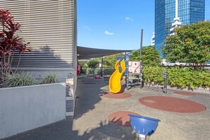 Ein Spielplatz mit einer gelben Rutsche und roten, runden Spielbereichen, umgeben von Grünflächen und einem modernen Gebäude.