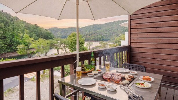 Un balcón con una mesa lista para una comida, una parrilla y vistas a un río y un bosque.