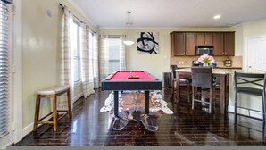 Une table de billard dans une pièce dotée d’un parquet en bois foncé, un coin cuisine avec des placards et un four à micro-ondes, et un coin repas avec des chaises et une table.