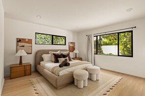 A modern bedroom with a large bed, two bedside tables, a window with a view of greenery, and artwork on the walls.