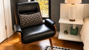 A modern living room with a leather armchair, a white side table with a lamp, and a view of a cityscape through large windows.