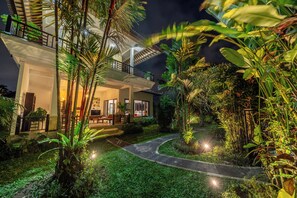 Una casa de dos pisos con un balcón, rodeada de exuberante vegetación y un camino de jardín bien iluminado.
