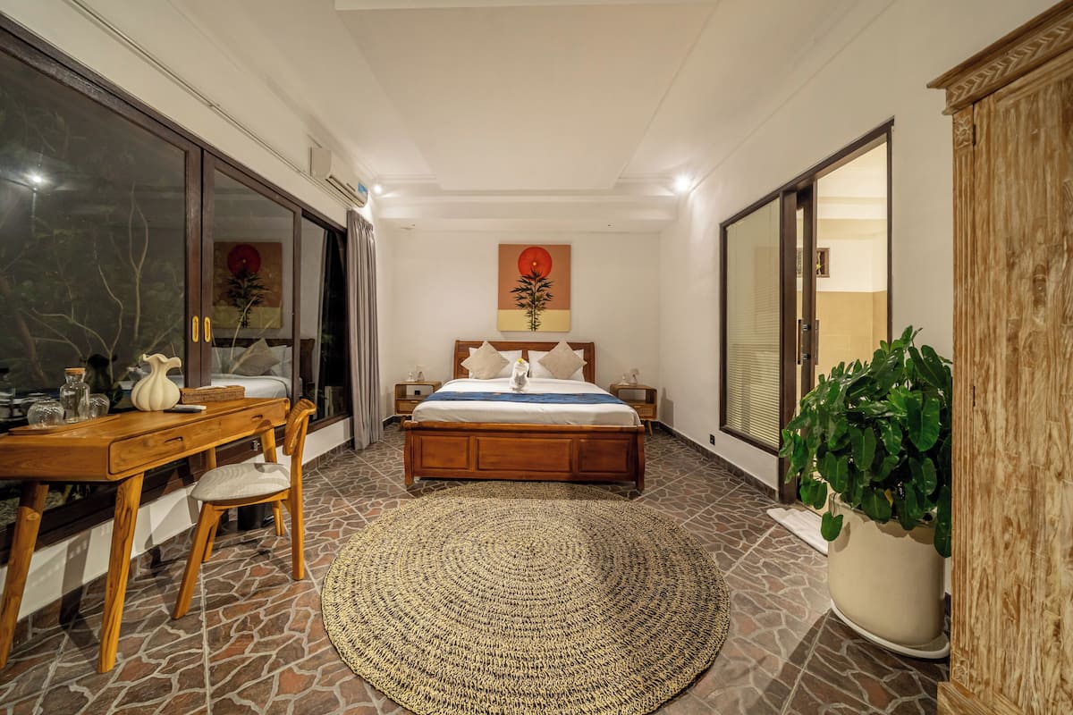 Un dormitorio con cama, escritorio, silla, una planta grande en maceta y un cuadro en la pared.