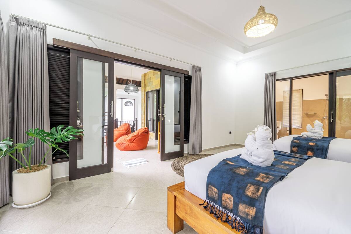 Una habitación de hotel con cama, planta en maceta, sillón de peluche y puerta corrediza de vidrio que da a un baño.