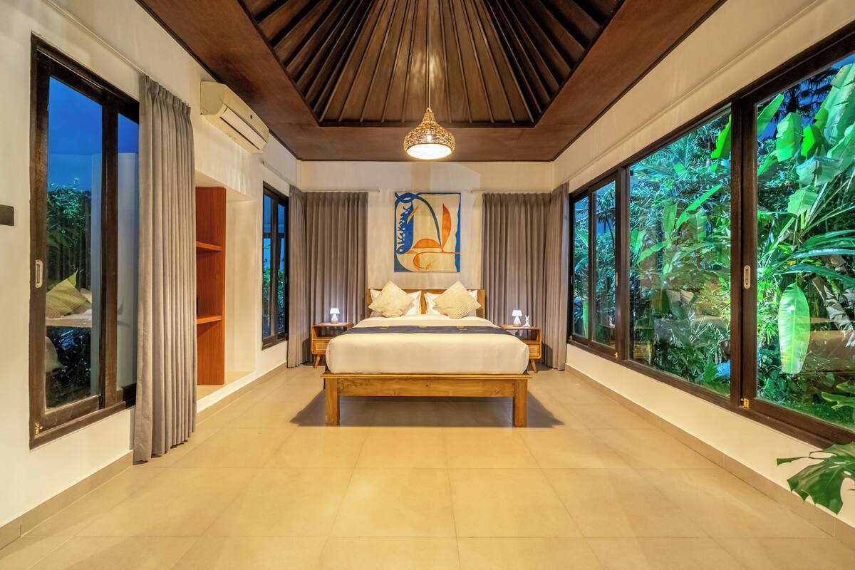 Un dormitorio con una cama grande, un ventilador de techo y vistas a zonas verdes a través de las ventanas.