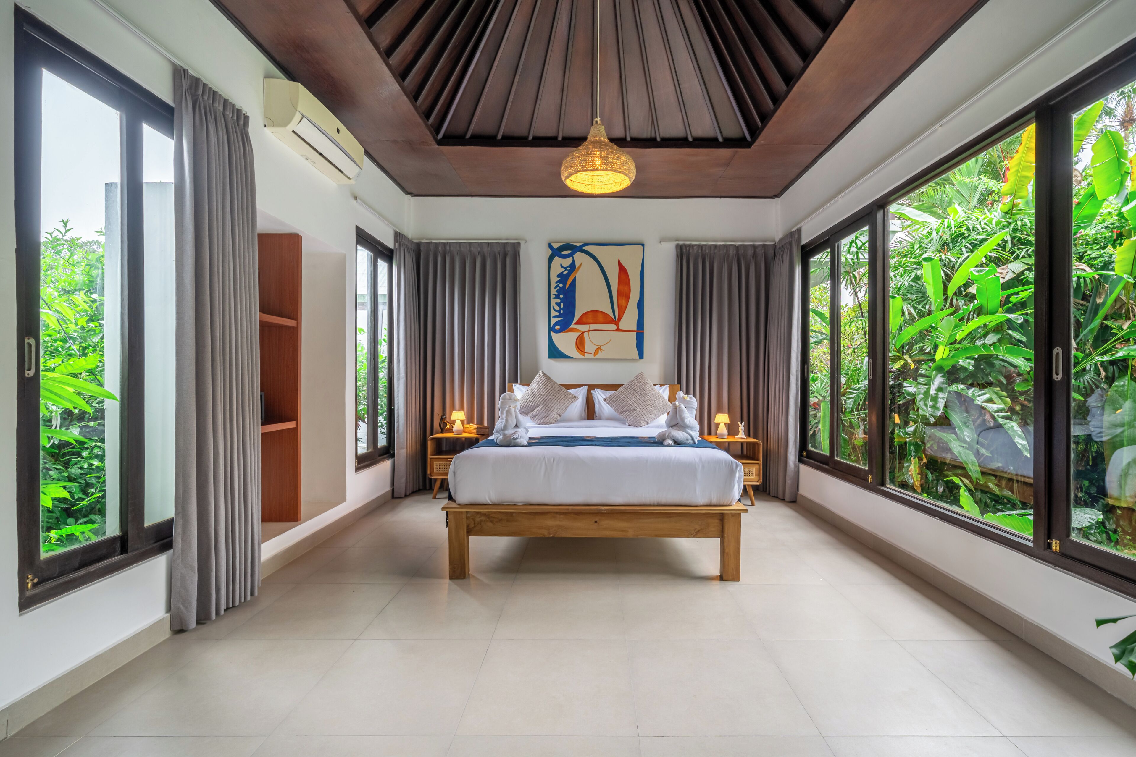 A spacious bedroom with a large bed, a ceiling fan, and a view of lush greenery through large windows.