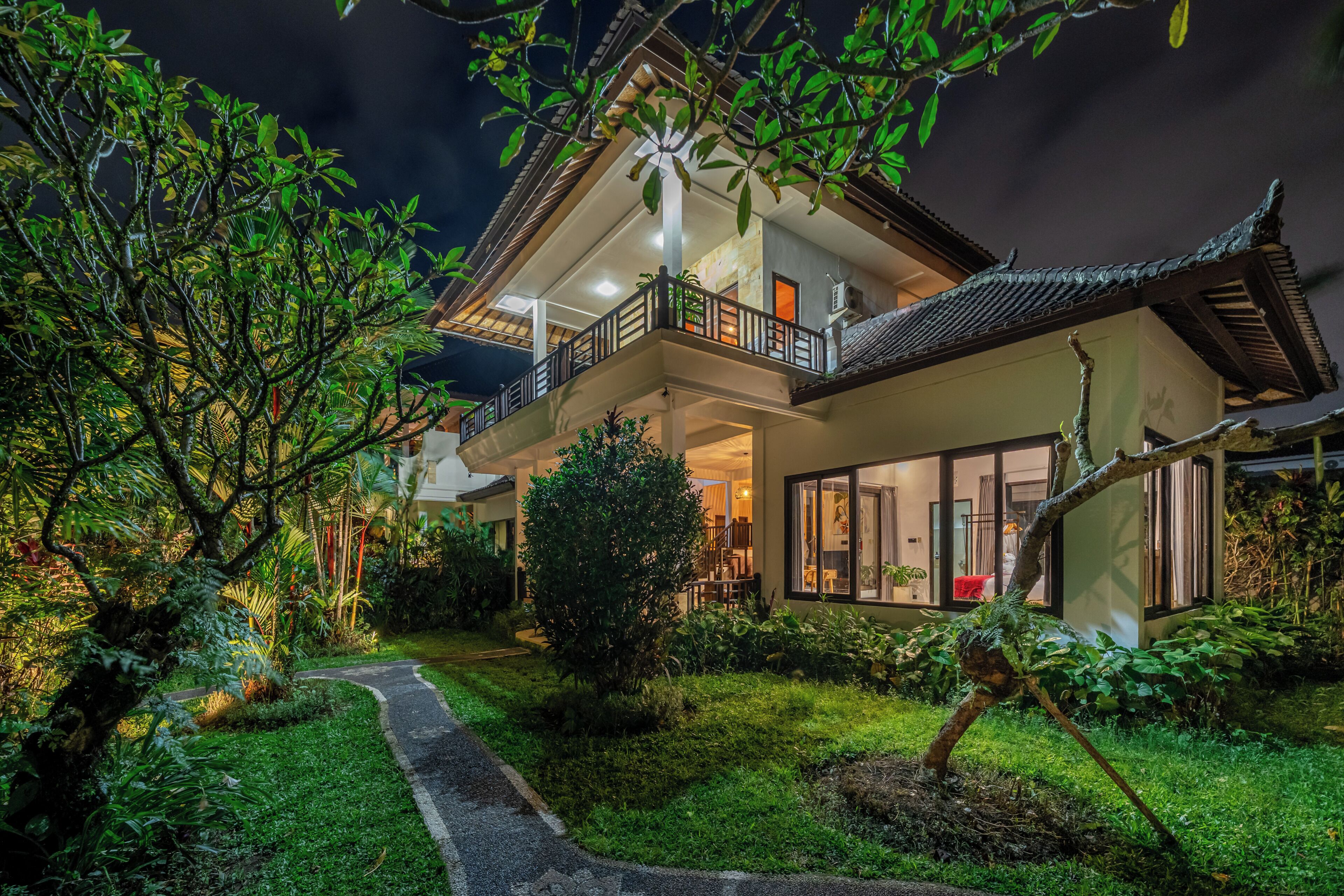Une maison à deux étages dotée d’un balcon, entourée d’une végétation luxuriante et accessible par un chemin menant à l’entrée.
