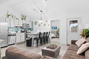 Eine moderne Küche mit Edelstahlgeräten, ein Essbereich mit Barhocker und ein Wohnzimmer mit Sofa und Hocker.
