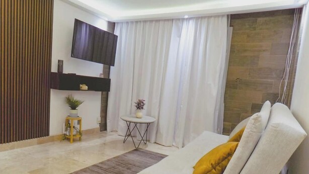 Ein Wohnzimmer mit einer Couch, einem kleinen runden Tisch, einem an der Wand befestigten Fernseher und einem Vorhang.