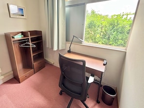 A room with a desk, chair, wardrobe, and a window with a view of greenery.