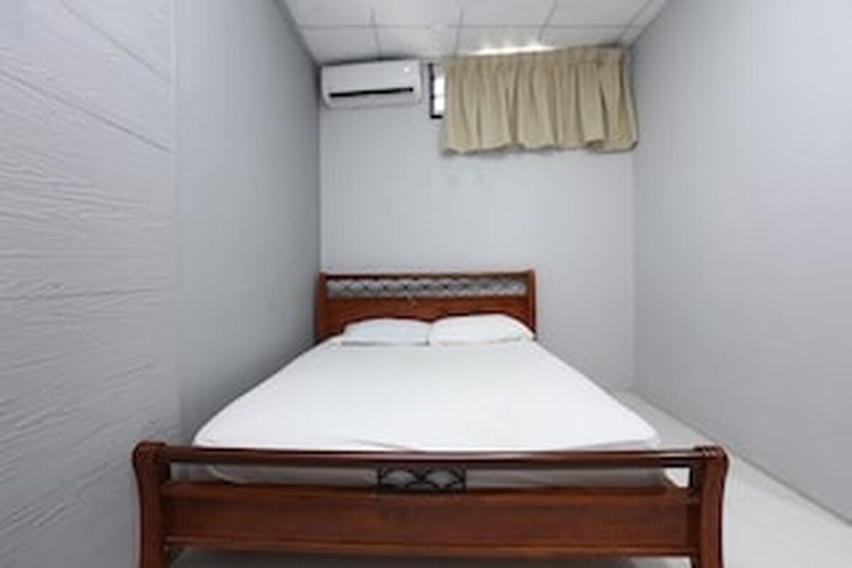 A small, narrow room with a single bed, a wooden headboard, and a white wall.