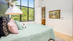 Ein Schlafzimmer mit großem Fenster, einem Bett mit vielen Kissen, einer Pendelleuchte und einem Bild an der Wand.