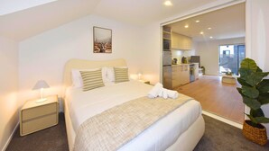 Un dormitorio moderno con una cama grande, lámparas de noche, un cuadro y vistas a la cocina y la sala de estar.
