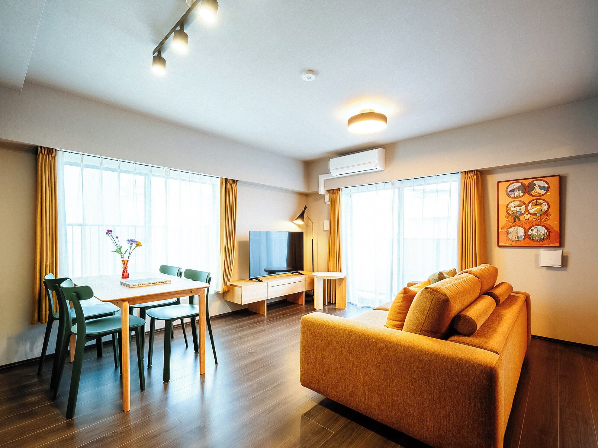 A hotel room with a wooden floor, a sofa, a TV, a desk, and a dining area with chairs and a table.
