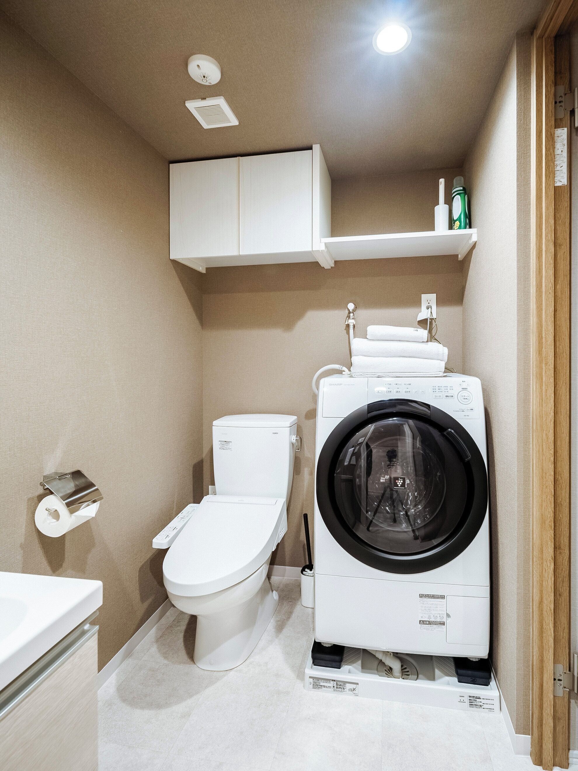 A compact bathroom with a toilet, washing machine, and a shelf above the toilet.