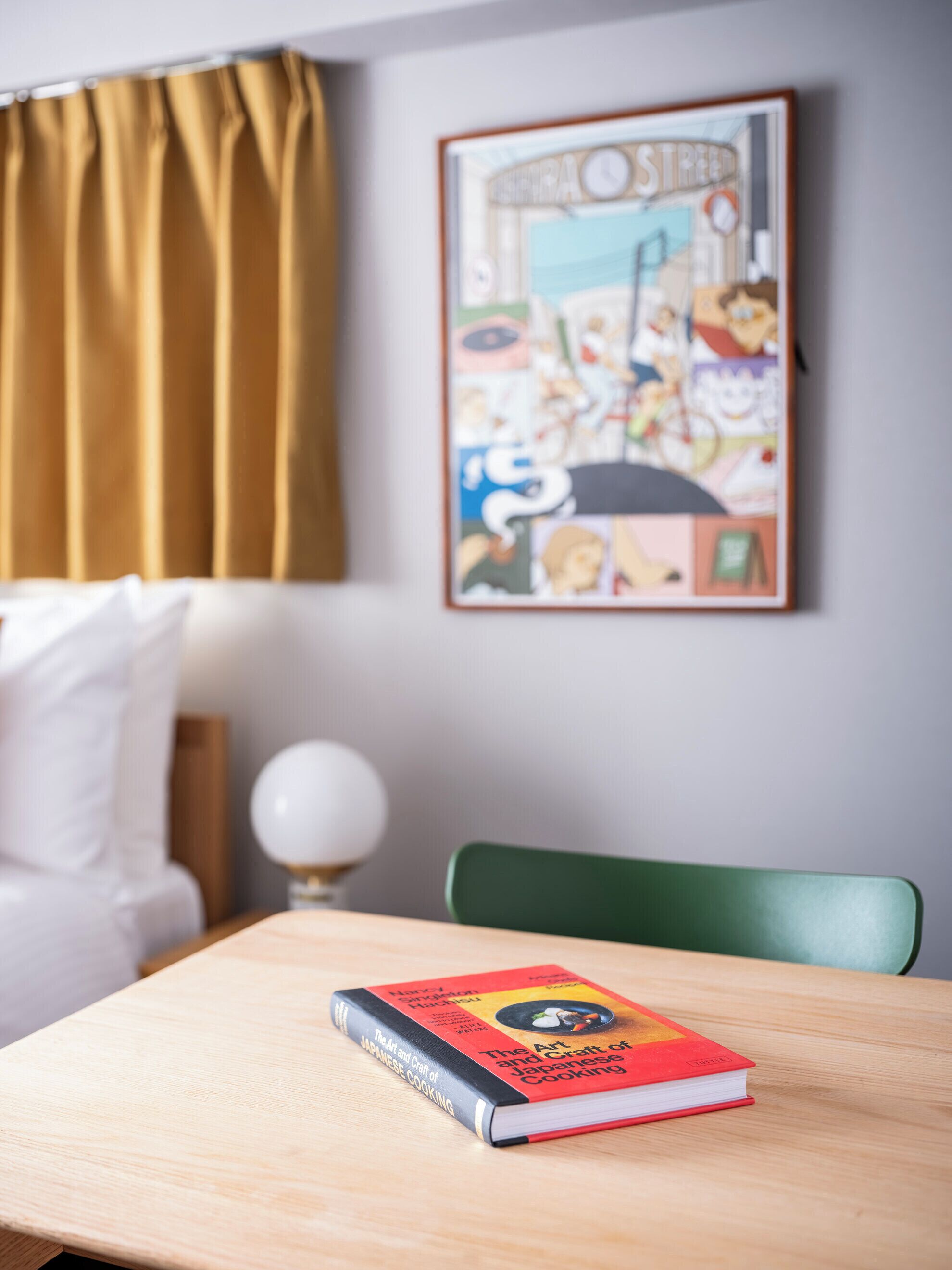 A book titled 'The Art and Craft of Japanese Cooking' on a wooden table in a room with a green chair, a white pillow, and a framed picture on the wall.
