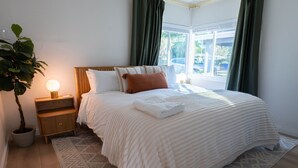 Een net opgemaakt bed met een witte deken en kussens, een bedlampje, een bloempot en uitzicht op het buiten via een raam met gordijnen.