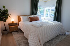Ein ordentlich bezogenes Bett mit weißer Decke und Kissen, eine Nachttischlampe, ein Blumentopf und ein Blick ins Freie durch ein Fenster mit Vorhängen.