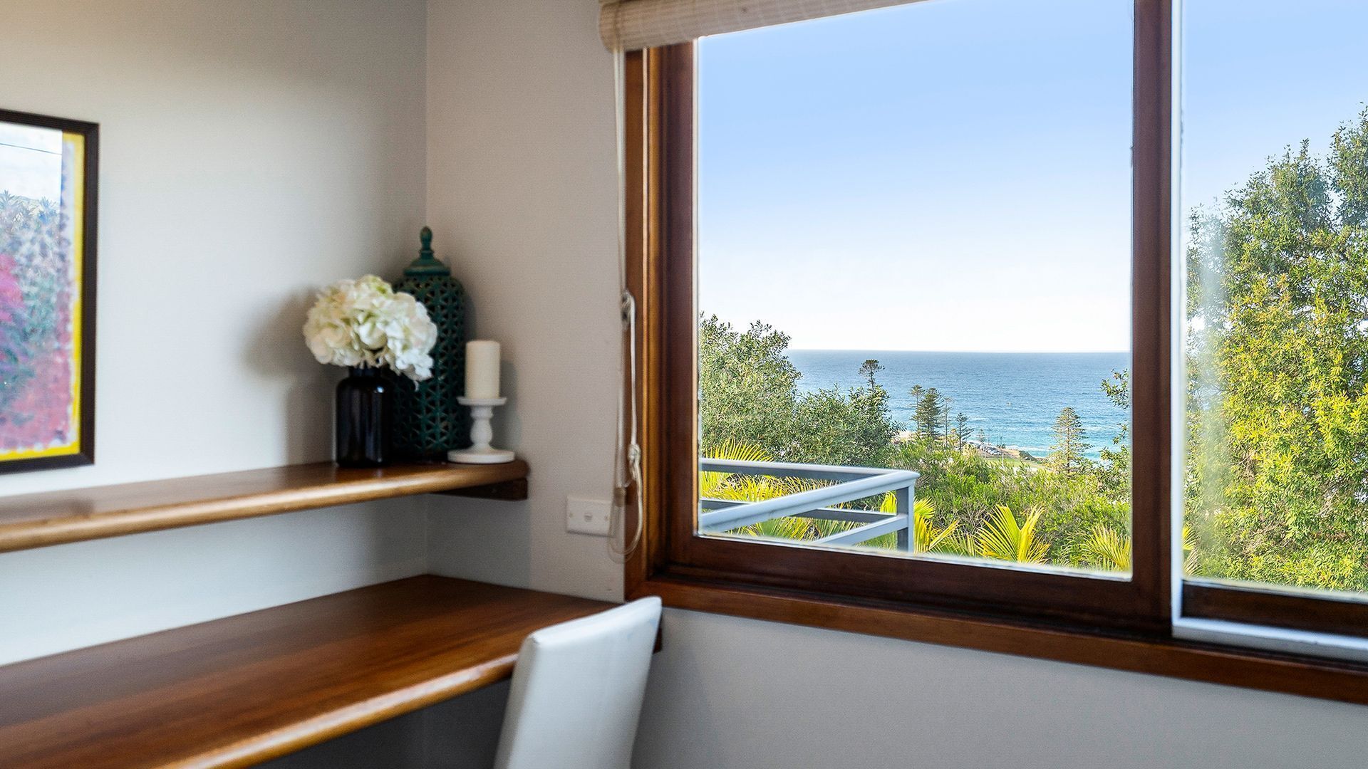 A room with a wooden desk, a window with a view of the ocean, and a shelf with a vase of flowers.