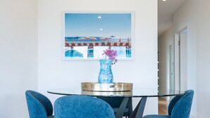Ein Essbereich mit einem runden Glastisch, blauen Stühlen, einer Vase mit violetten Blumen und einem Bilderrahmen mit einer Strandansicht.